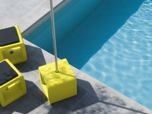 drie exemplaren van cube aan een zwembad, één gebruikt als parasolvoet, twee als pouf met een kussentje erop