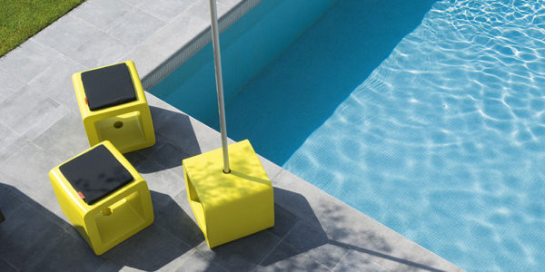 drie exemplaren van cube aan een zwembad, één gebruikt als parasolvoet, twee als pouf met een kussentje erop